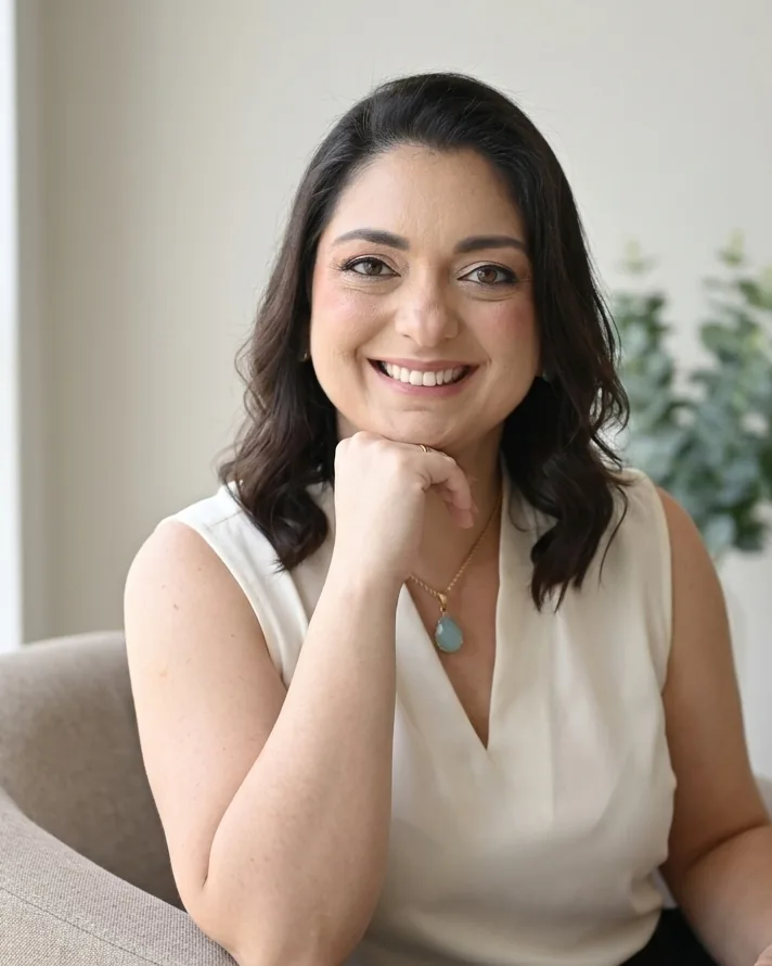 Débora Aires sorrindo, sentada em uma poltrona clara com um livro na mão